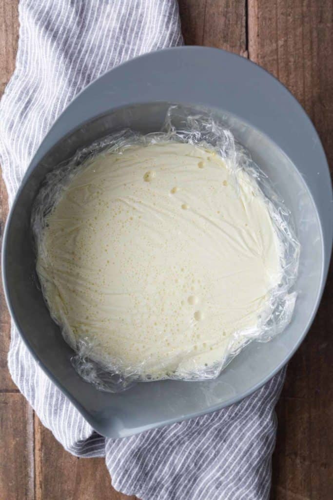 A mixing bowl lined with plastic wrap contains a smooth, light yellow mixture, resting on a striped cloth on a wooden surface.