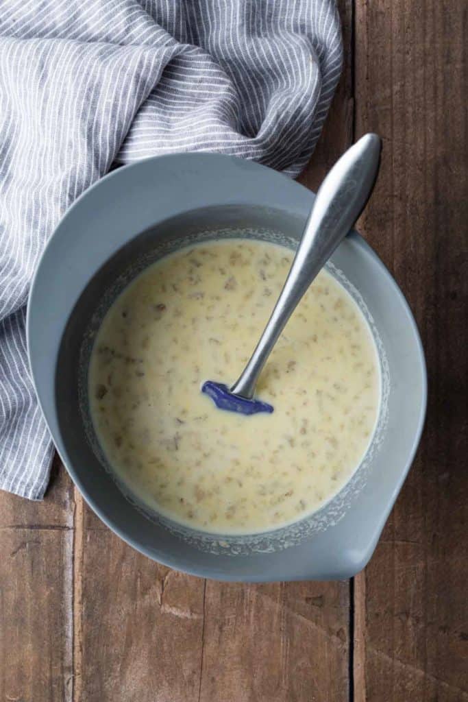 A mixing bowl with a spoon in a pale yellow liquid mixture containing small bits, sitting on a wooden surface next to a striped cloth.