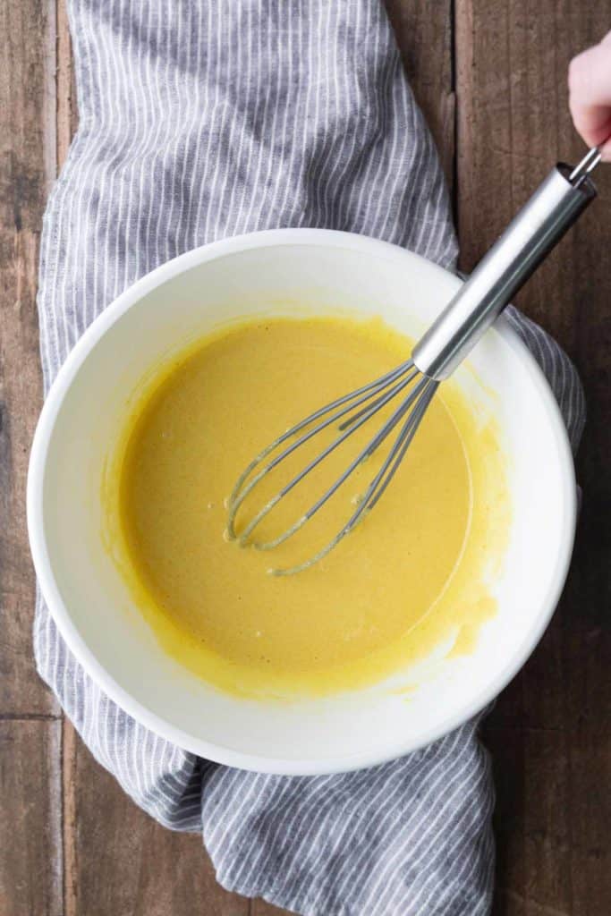 A metal whisk rests in a white bowl containing yellow batter on a gray striped cloth, set on a wooden surface.