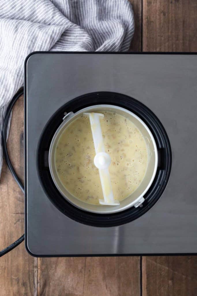 Overhead view of an ice cream maker with a creamy mixture inside, sitting on a wooden surface next to a striped cloth.