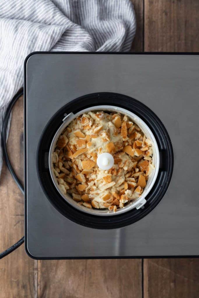Overhead view of a food processor containing a mixture topped with crushed crackers, placed on a wooden surface next to a striped cloth.