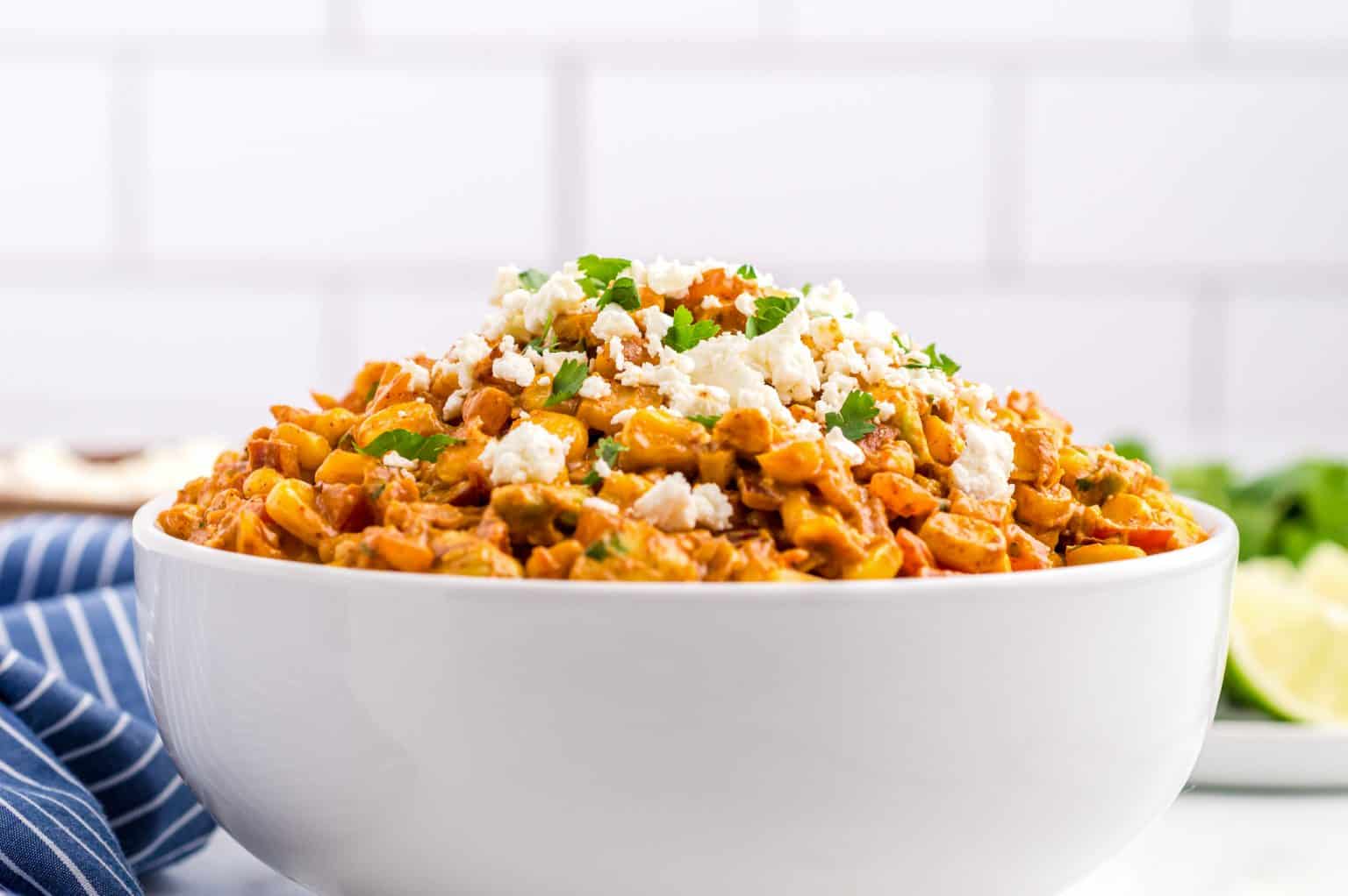 A white bowl filled with Mexican street corn salad topped with crumbled cheese and chopped cilantro, with a striped napkin and lime wedges in the background.