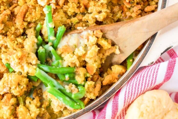 A wooden spoon rests in a dish of green bean casserole topped with crumbled stuffing, next to a red and white striped towel and a biscuit.