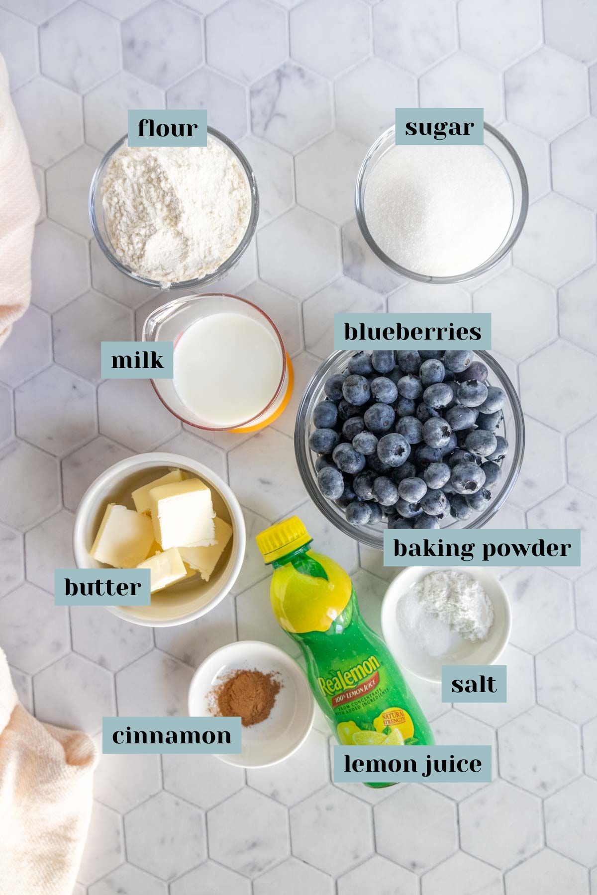 Top-down view of labeled bowls containing flour, sugar, blueberries, milk, butter, baking powder, cinnamon, salt, and a bottle of lemon juice on a white hexagonal surface.