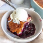 A bowl with a serving of blueberry cobbler topped with a scoop of vanilla ice cream; fresh blueberries are in the background.
