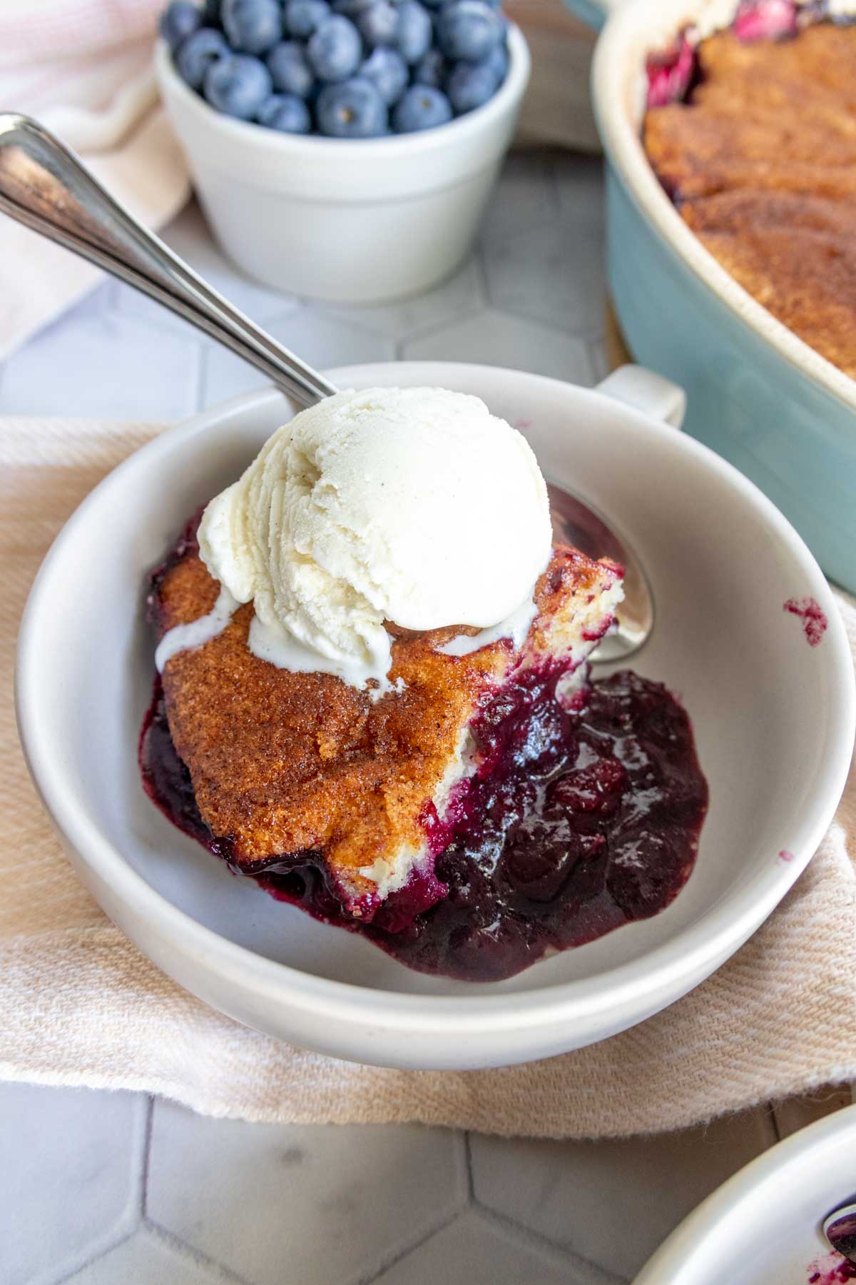 A bowl with a serving of blueberry cobbler topped with a scoop of vanilla ice cream; fresh blueberries are in the background.