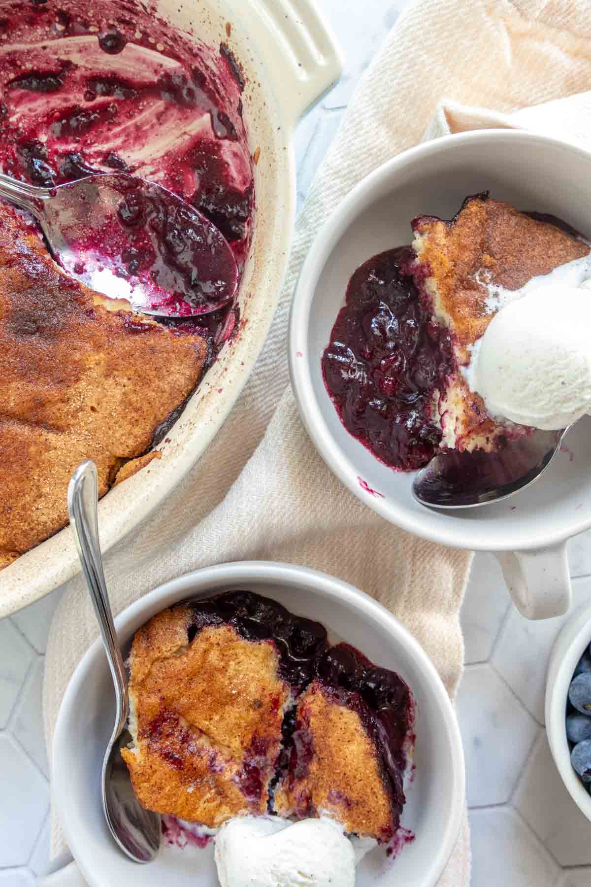 Overhead view of two bowls of blueberry cobbler with vanilla ice cream, next to a baking dish containing the remaining cobbler.