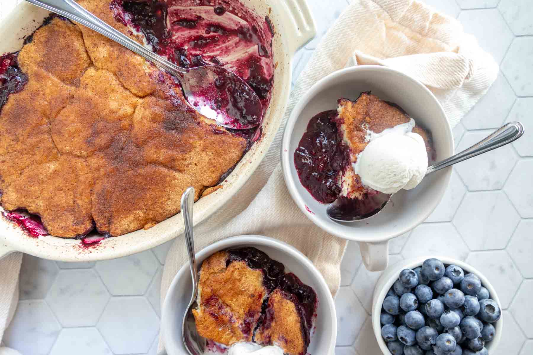 Top view of a blueberry cobbler in a baking dish with two bowls of cobbler topped with vanilla ice cream, and a bowl of fresh blueberries on the side.