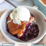 A bowl of blueberry cobbler topped with a scoop of vanilla ice cream, with a spoon beside it and fresh blueberries in the background.
