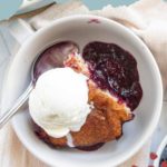 A bowl of blueberry cobbler topped with a scoop of vanilla ice cream, served with a spoon on a light cloth.