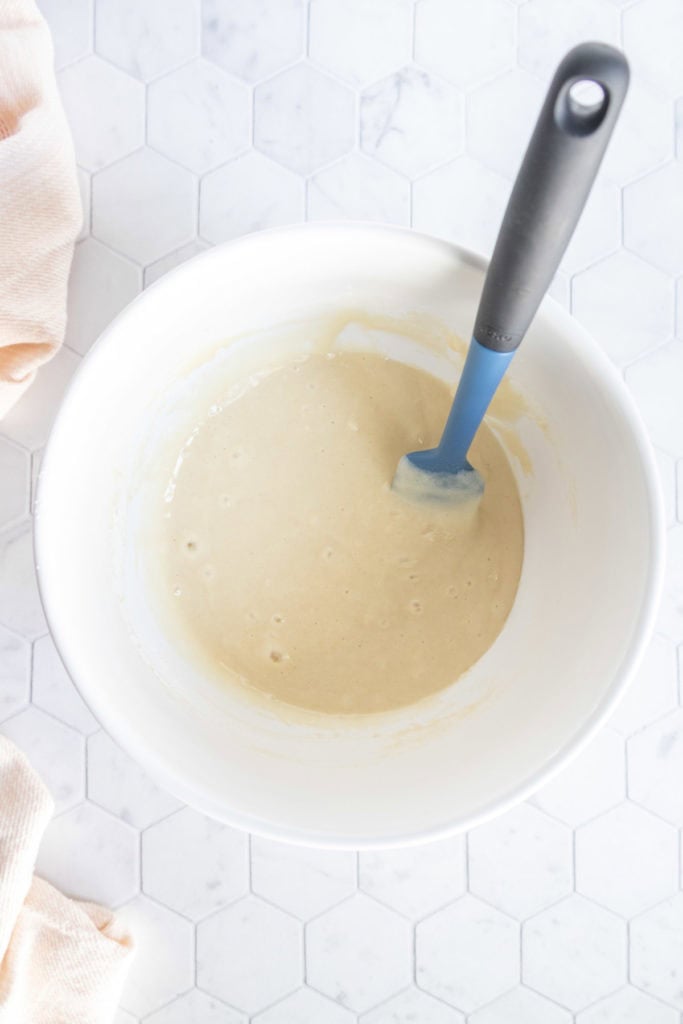 A white mixing bowl with light-colored batter and a blue spatula sits on a white hexagonal tile surface.