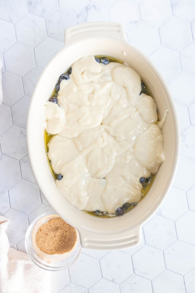 An oval baking dish filled with unbaked batter and blueberries, next to a small container of brown sugar on a white hexagon-tiled surface.