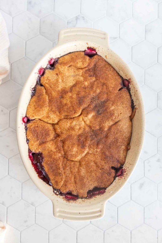 A baked cobbler with a golden-brown crust and bubbling fruit filling is shown in an oval ceramic dish on a hexagon-tiled surface.