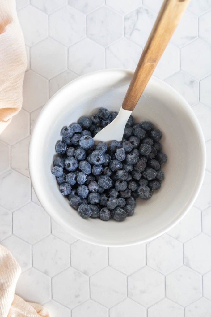 A white bowl filled with blueberries and a white spatula with a wooden handle, placed on a white hexagon tile surface.