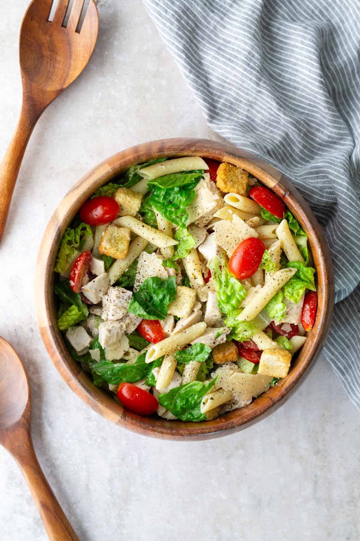 A wooden bowl filled with pasta salad containing penne, chopped lettuce, cherry tomatoes, croutons, and chunks of chicken, next to wooden utensils and a striped cloth.