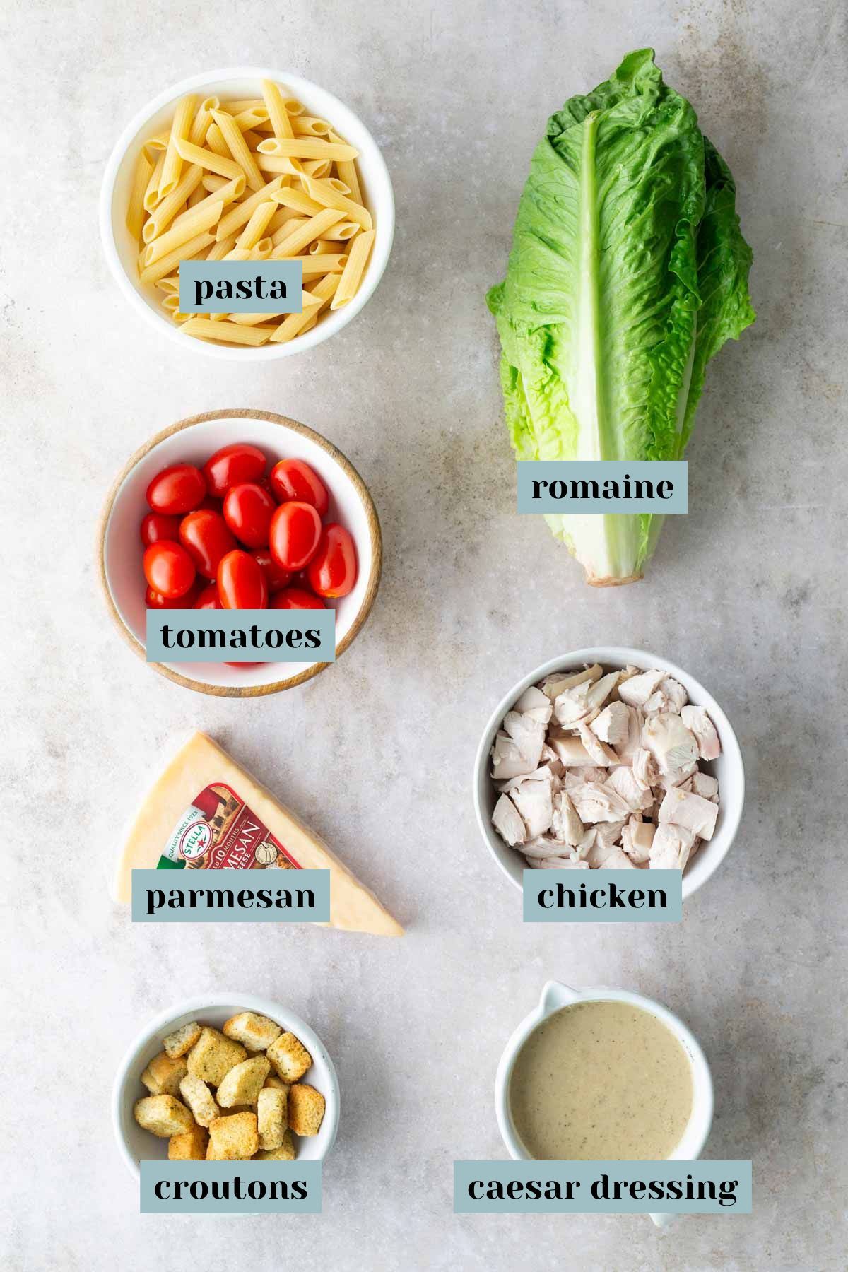 Flat lay of ingredients for a chicken Caesar pasta salad: pasta, romaine lettuce, cherry tomatoes, parmesan cheese, chicken, croutons, and Caesar dressing, each labeled.