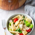 A bowl of pasta salad with penne, romaine lettuce, cherry tomatoes, croutons, chicken, and parmesan, with a fork beside it and a larger wooden salad bowl in the background.