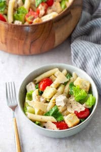 A bowl of pasta salad with penne, romaine lettuce, cherry tomatoes, croutons, chicken, and parmesan, with a fork beside it and a larger wooden salad bowl in the background.