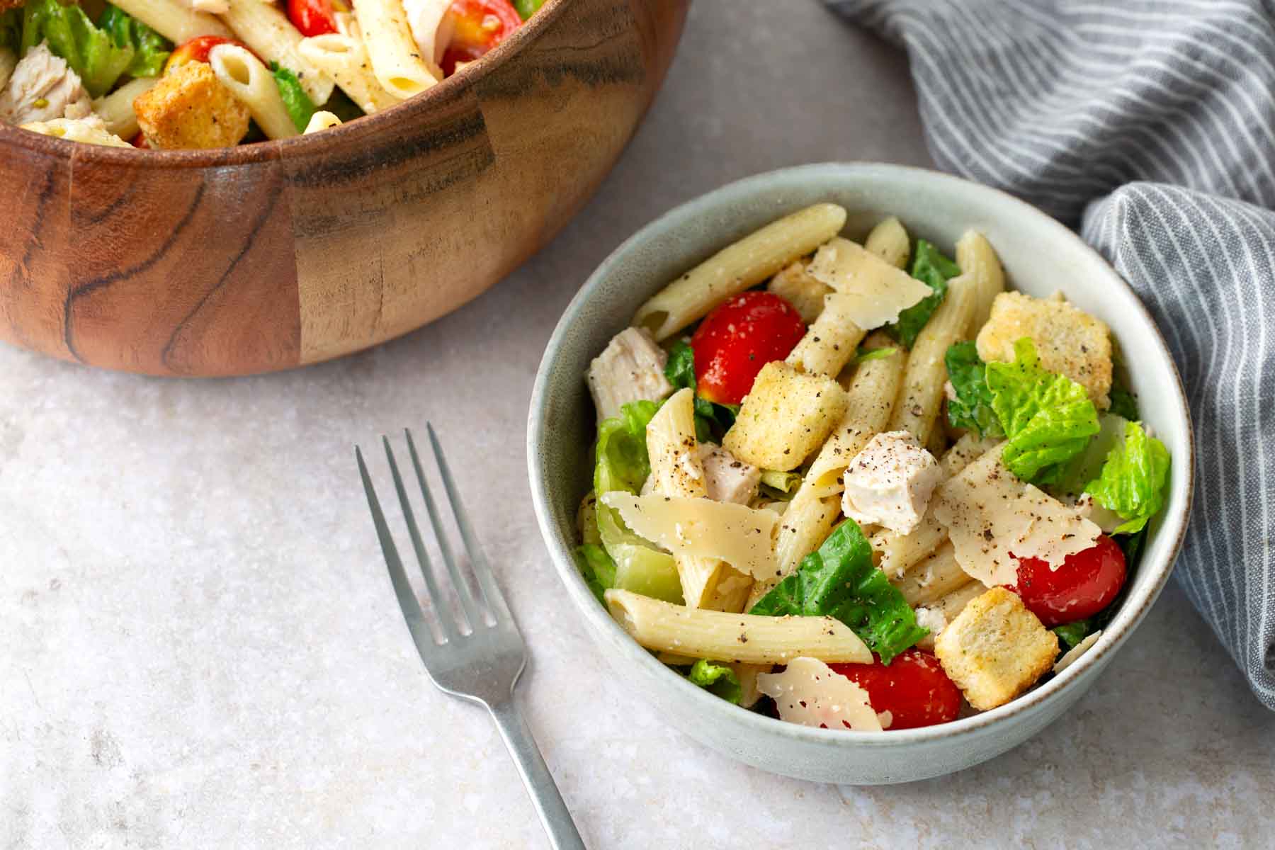 A bowl of pasta salad with penne, cherry tomatoes, lettuce, croutons, shaved cheese, and black pepper, next to a fork and a larger salad bowl.