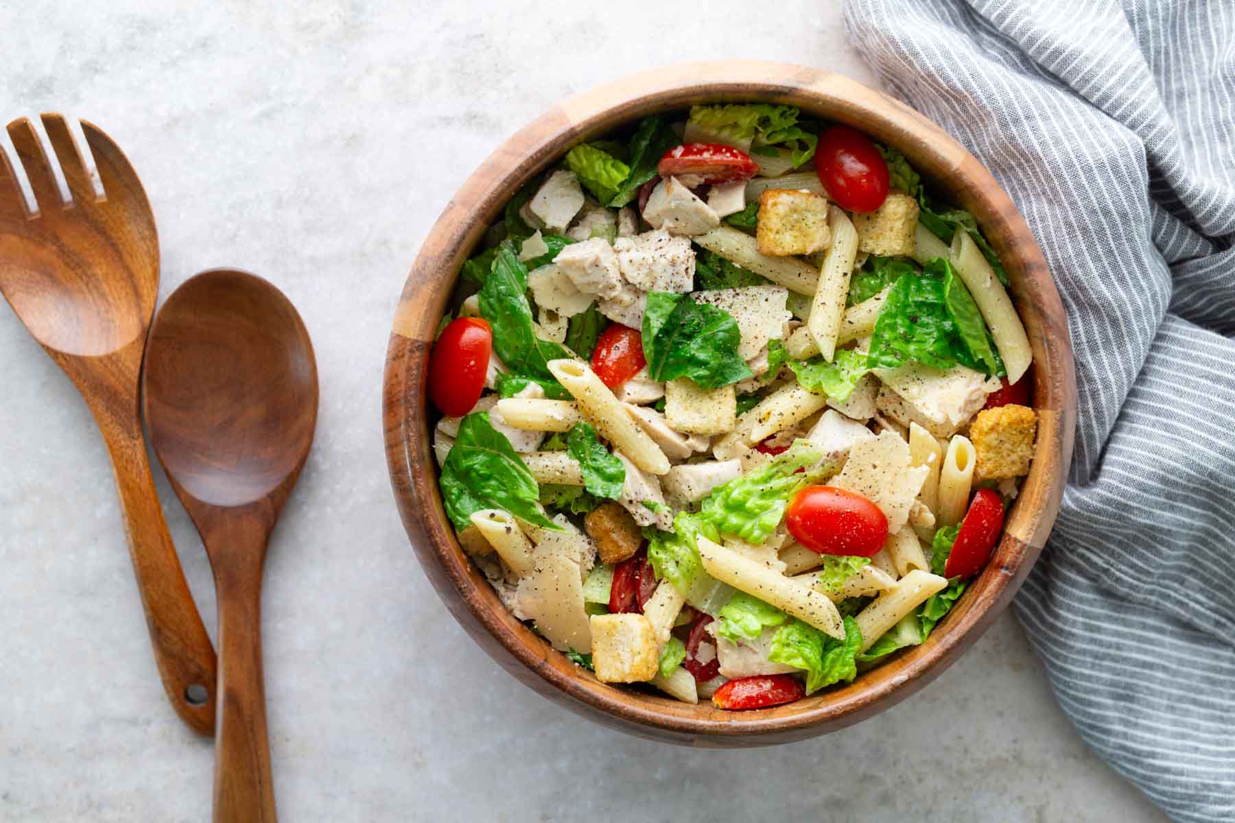 A wooden bowl filled with pasta salad containing penne, lettuce, cherry tomatoes, croutons, and chicken pieces, with wooden salad servers and a striped cloth nearby.