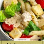 Bowl of chicken Caesar pasta salad with penne, lettuce, cherry tomatoes, croutons, parmesan slices, and black pepper.