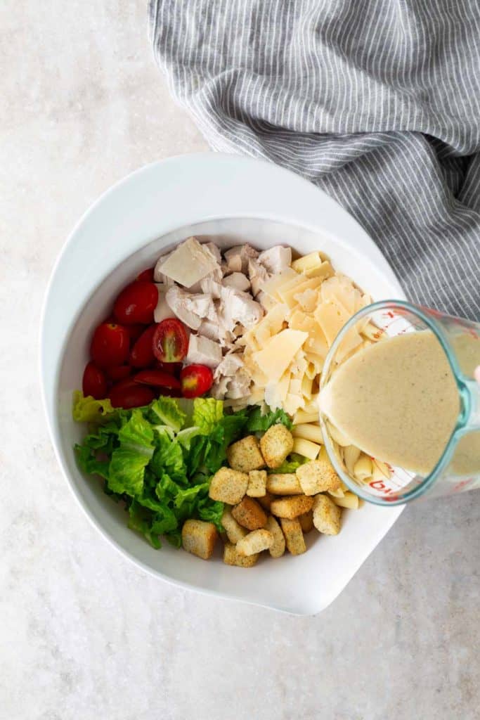 A white bowl containing lettuce, cherry tomatoes, cooked chicken, shredded cheese, croutons, and Caesar dressing being poured in.