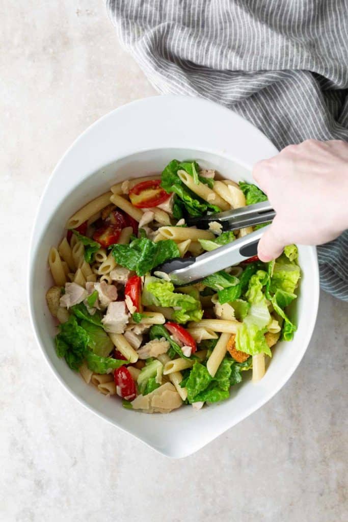 A hand using tongs to toss a salad with penne pasta, lettuce, tomatoes, olives, and chicken in a white bowl, with a striped cloth nearby.