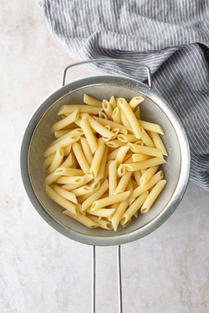 Cooked penne pasta draining in a metal colander, placed on a light countertop next to a gray and white striped cloth.