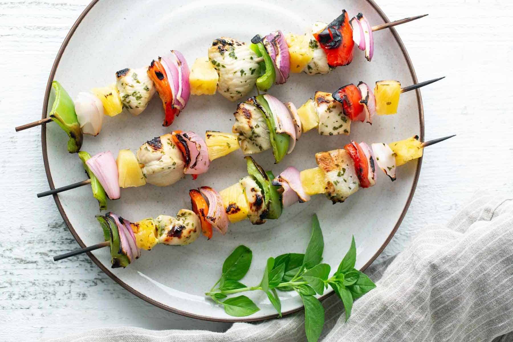 Three grilled chicken and vegetable kebabs on a white plate with a sprig of fresh basil and a striped cloth in the background.