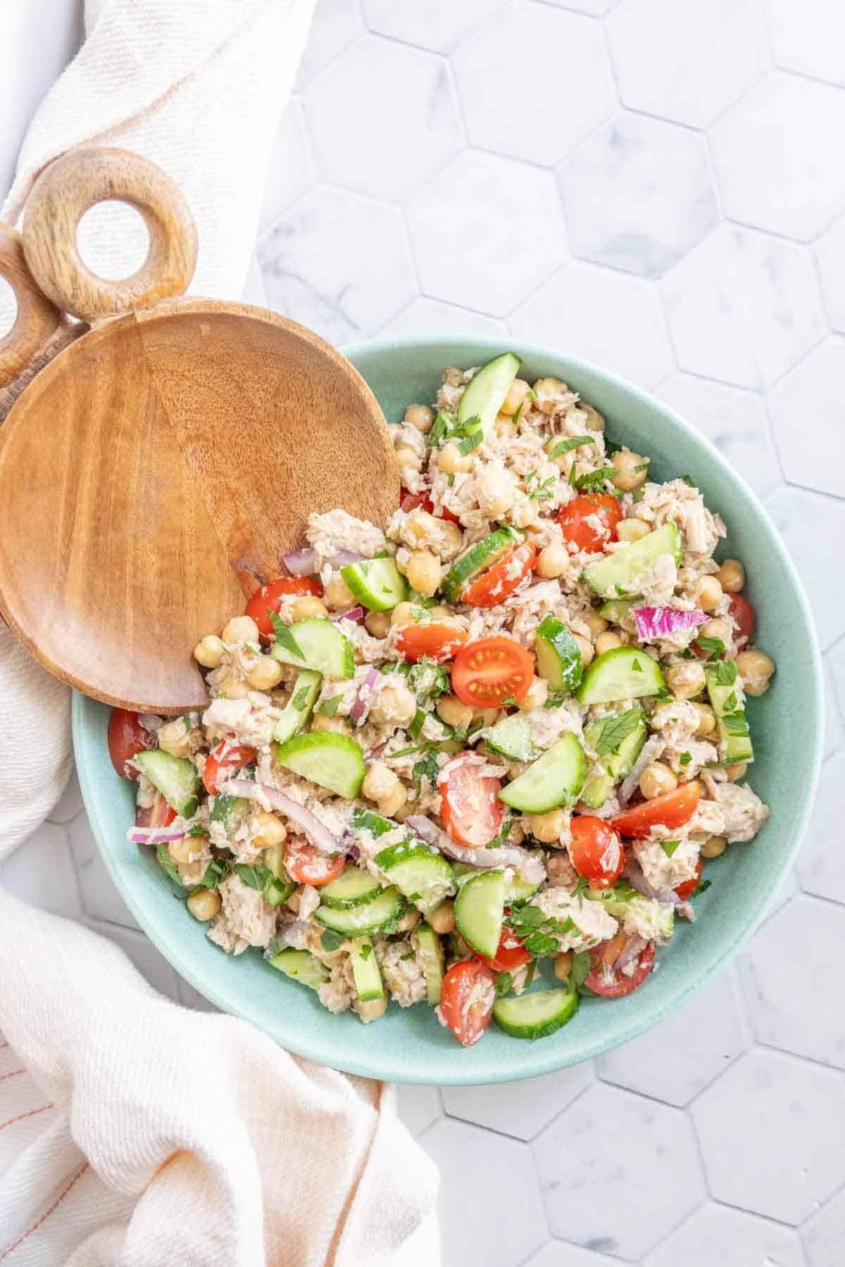 A bowl of salad with chickpeas, cherry tomatoes, cucumber, red onion, and herbs, with a wooden serving spoon on the side.
