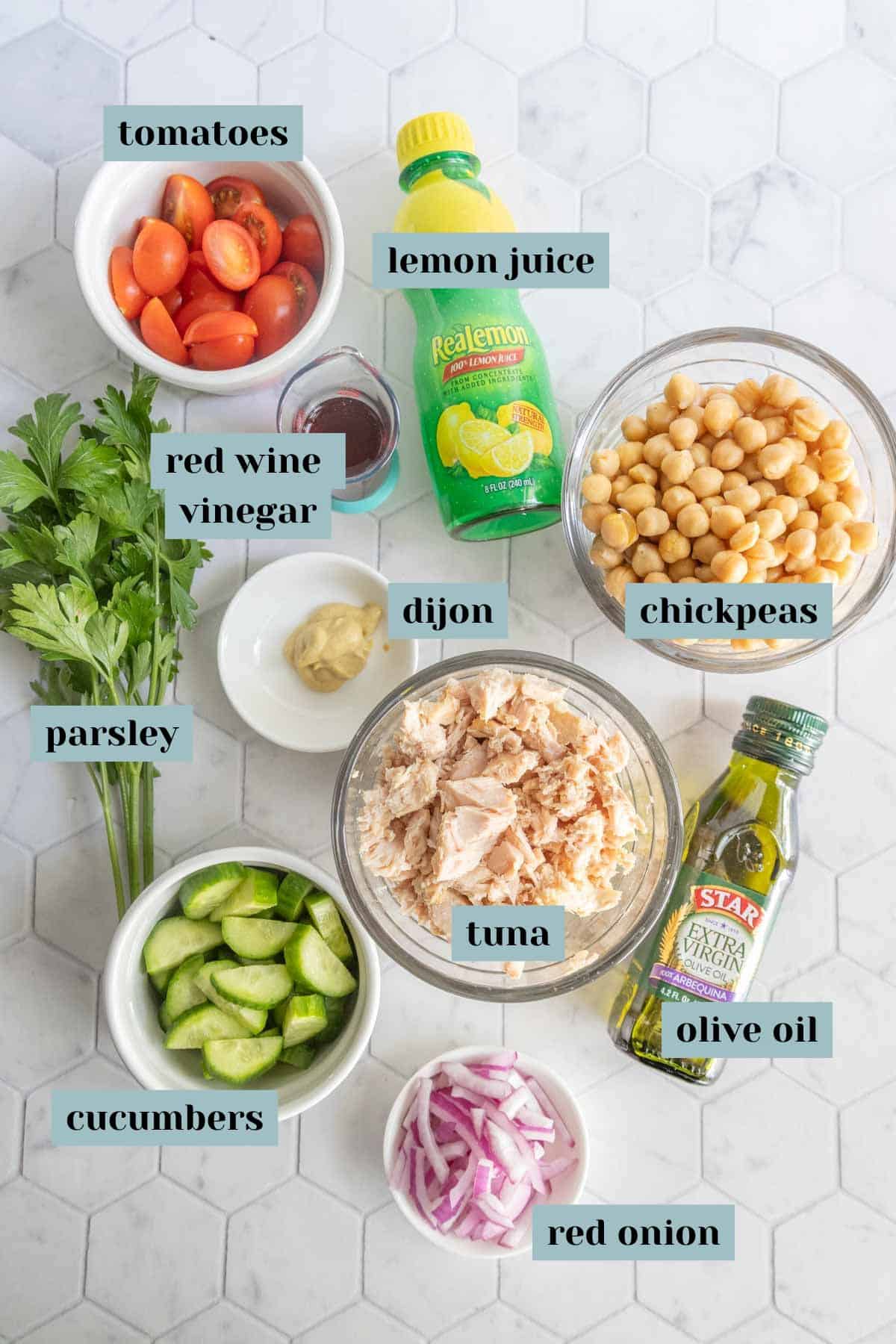 Bowls and bottles containing cherry tomatoes, lemon juice, red wine vinegar, chickpeas, parsley, cucumbers, dijon mustard, tuna, olive oil, and sliced red onion on a white surface.