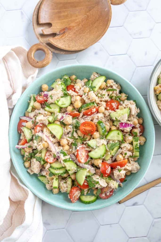 A bowl of salad containing chickpeas, cherry tomatoes, cucumber, red onion, tuna, and mixed greens on a white hexagon-tiled surface.