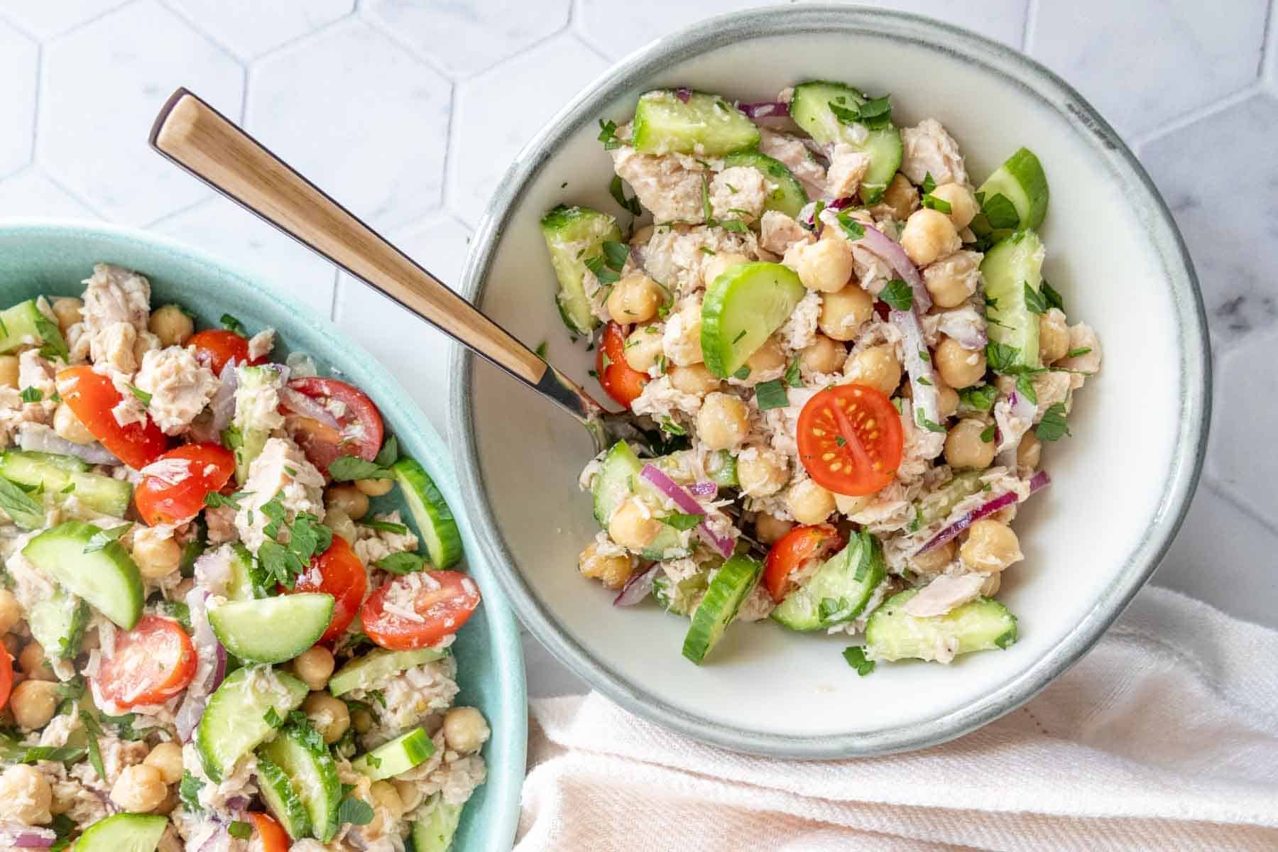 A bowl of chickpea salad with cherry tomatoes, cucumber, red onion, and herbs, served with a fork; a larger bowl of the same salad is in the background.