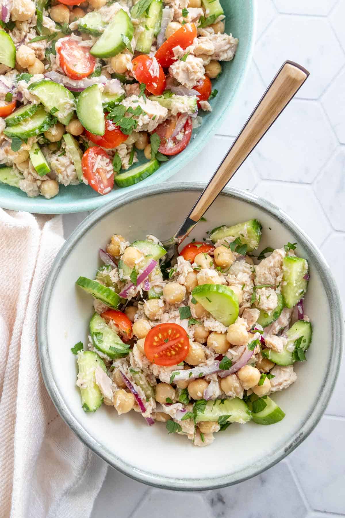 A bowl of chickpea salad with cherry tomatoes, cucumber, red onion, and herbs, served with a fork; a larger bowl of the same salad is in the background.