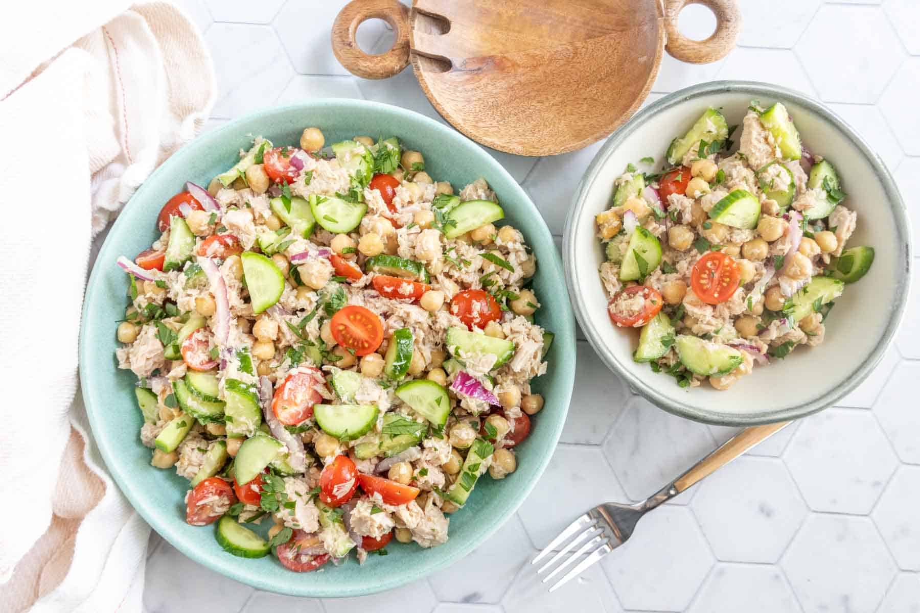 Two bowls of chickpea salad with tuna, cherry tomatoes, cucumber, red onion, and herbs on a white hexagonal tile surface, with a fork and wooden serving utensils nearby.
