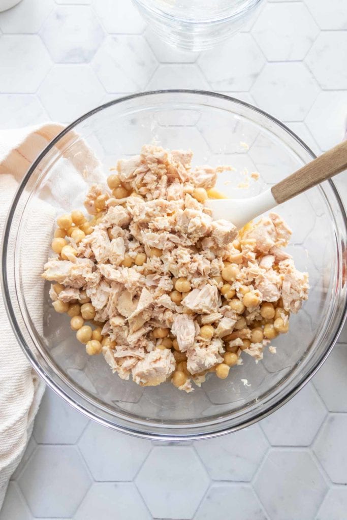 A glass bowl filled with chunks of tuna and chickpeas, being mixed with a white spatula on a white hexagon-tiled surface.
