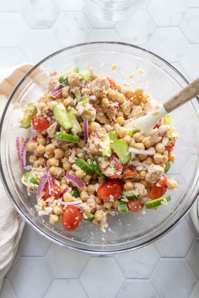 A glass bowl containing a mixed salad with chickpeas, cherry tomatoes, cucumber, red onion, tuna, and crumbled cheese, with a spatula resting inside the bowl.