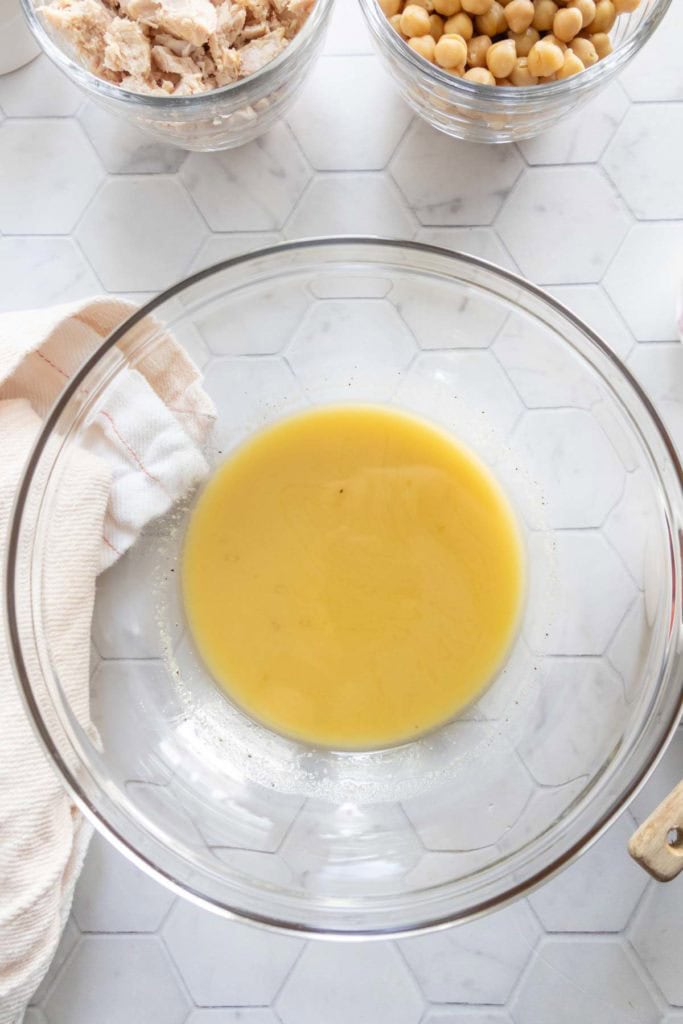 A glass mixing bowl with a small amount of yellow liquid sits on a white hexagon-tiled surface, with chickpeas, shredded chicken, and a towel nearby.