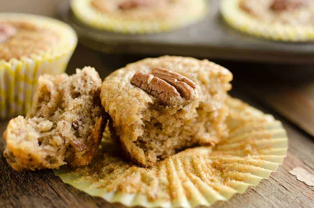 A pecan muffin split in half sits on a yellow muffin liner, with more muffins in the background.
