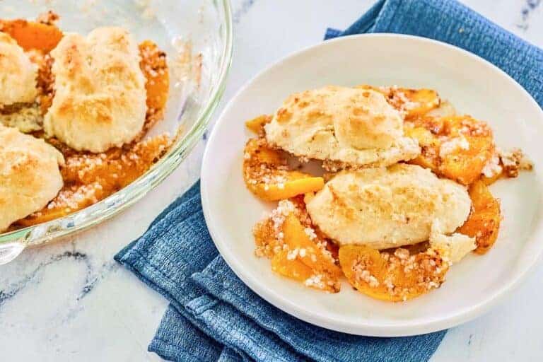 A serving of peach cobbler with biscuit topping is plated on a white dish, next to a glass baking dish with more cobbler, all on a blue cloth.