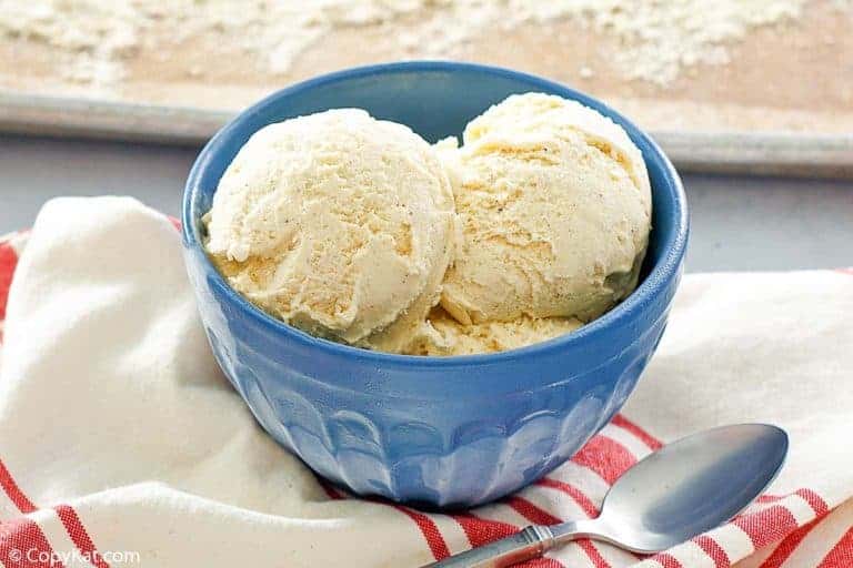 A blue bowl with two scoops of vanilla ice cream sits on a red and white striped cloth next to a metal spoon.