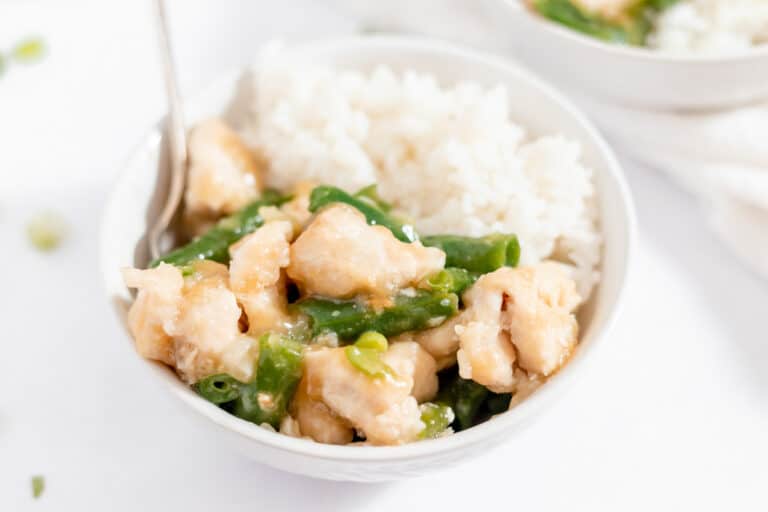 A bowl of white rice served with pieces of cooked chicken and green beans in a light sauce, with a fork on the side.