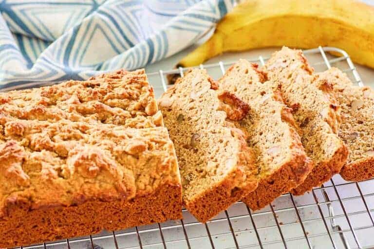 Sliced banana bread on a cooling rack with a banana and blue-and-white cloth in the background.