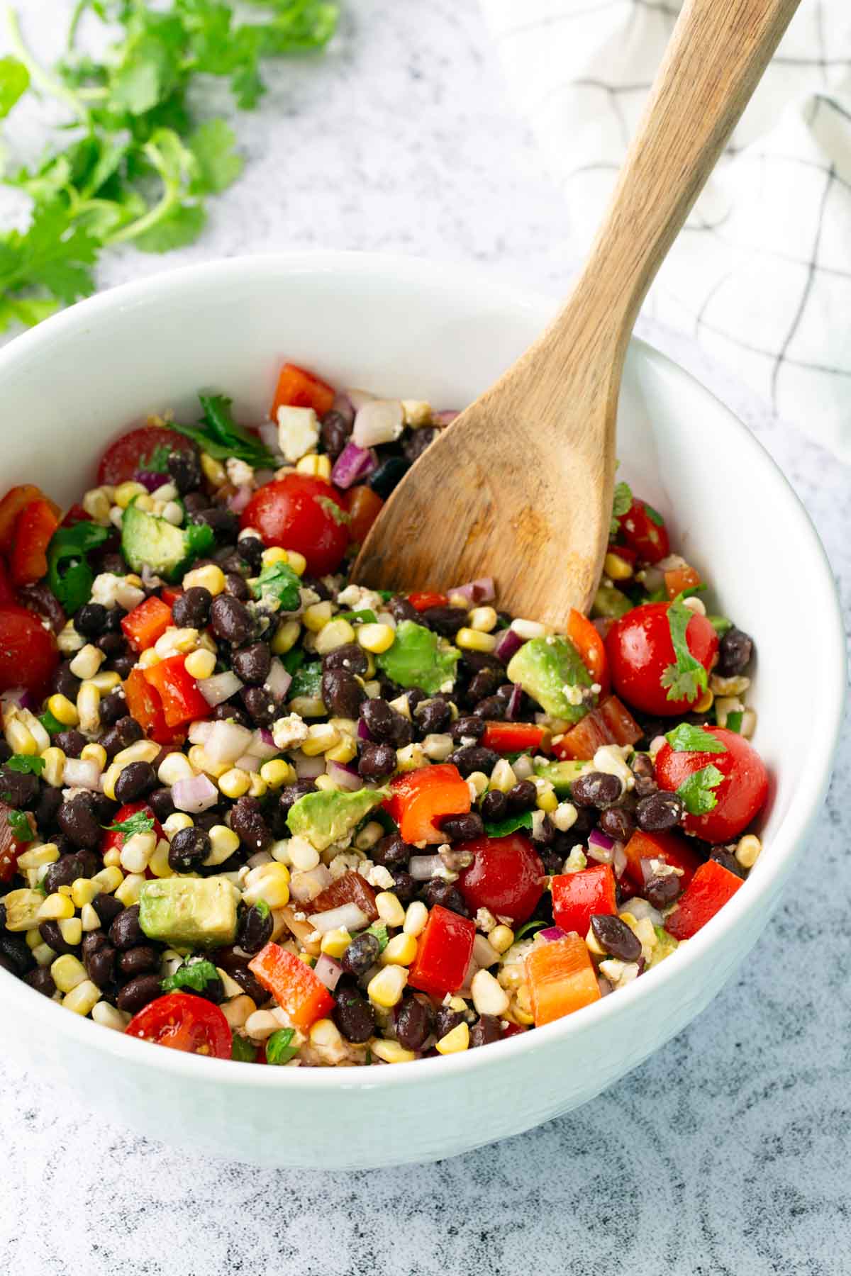 A white bowl filled with a colorful salad of black beans, corn, cherry tomatoes, red bell pepper, avocado, and cilantro, with a wooden spoon for mixing.