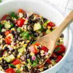 A white bowl filled with black bean salad containing corn, cherry tomatoes, red onion, avocado, and cilantro, with a wooden spoon resting inside.