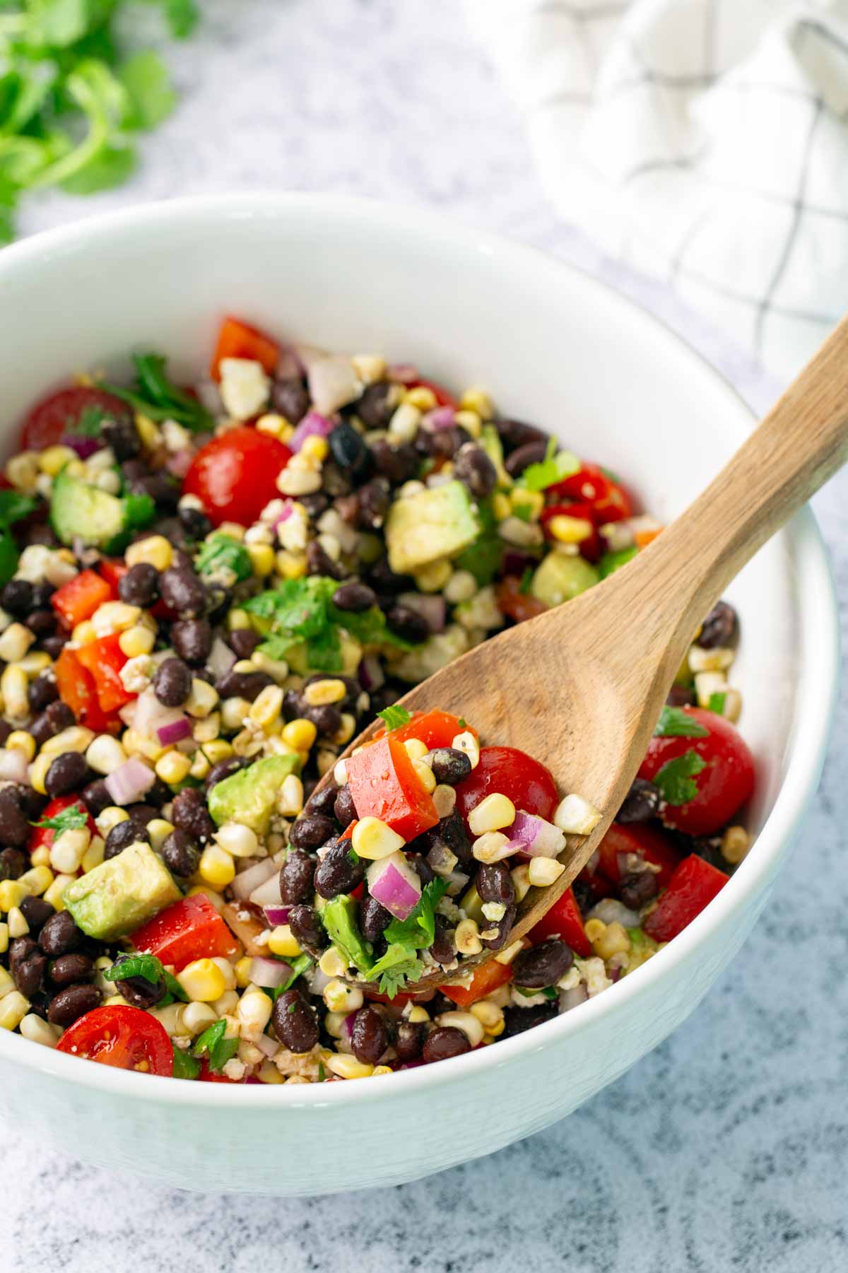 A white bowl filled with black bean salad containing corn, cherry tomatoes, red onion, avocado, and cilantro, with a wooden spoon resting inside.