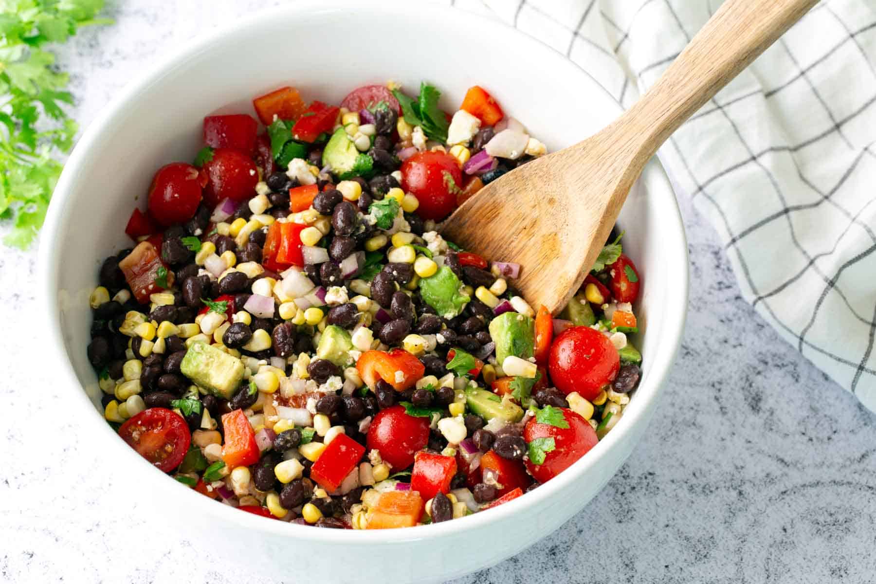 A white bowl filled with a black bean salad containing corn, cherry tomatoes, red bell pepper, avocado, and chopped onions, with a wooden spoon resting inside.