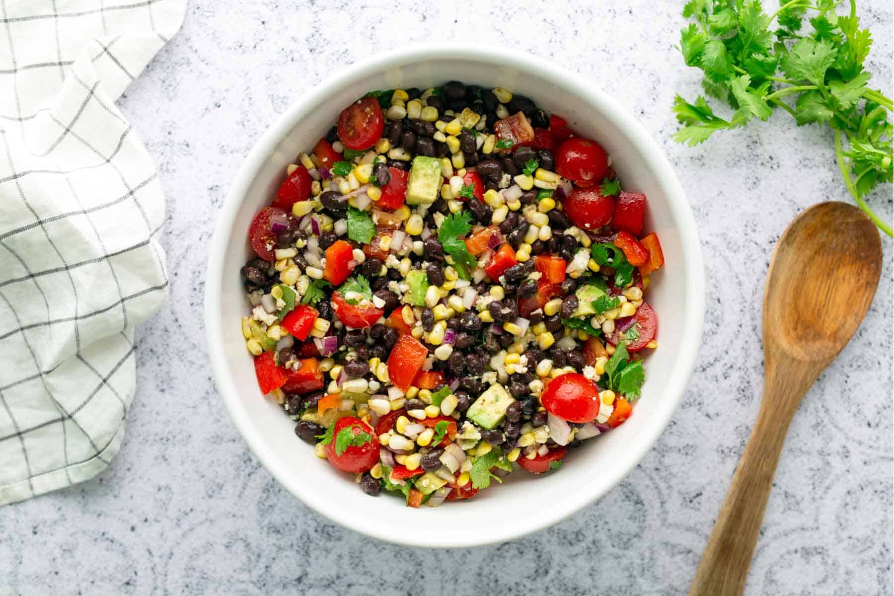 A bowl of black bean and corn salad with chopped tomatoes, red bell pepper, avocado, and cilantro on a light surface, next to a wooden spoon, napkin, and fresh cilantro.