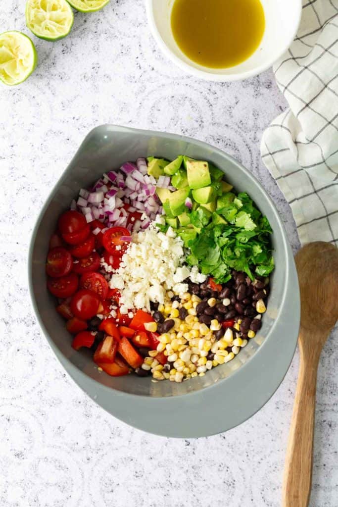 A gray bowl contains diced red onion, avocado, cherry tomatoes, cilantro, black beans, corn, chopped red pepper, and crumbled cheese. A bowl of dressing and wooden spoon are nearby.
