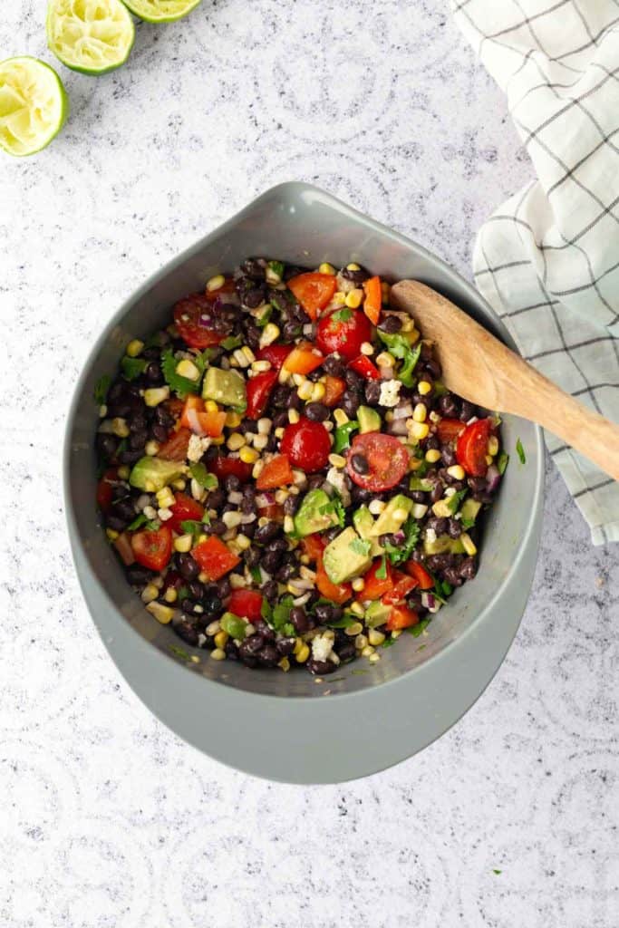 A gray bowl filled with a colorful salad of black beans, corn, tomatoes, avocado, and herbs sits on a light surface with a wooden spoon and a cloth nearby. Squeezed limes are in the corner.